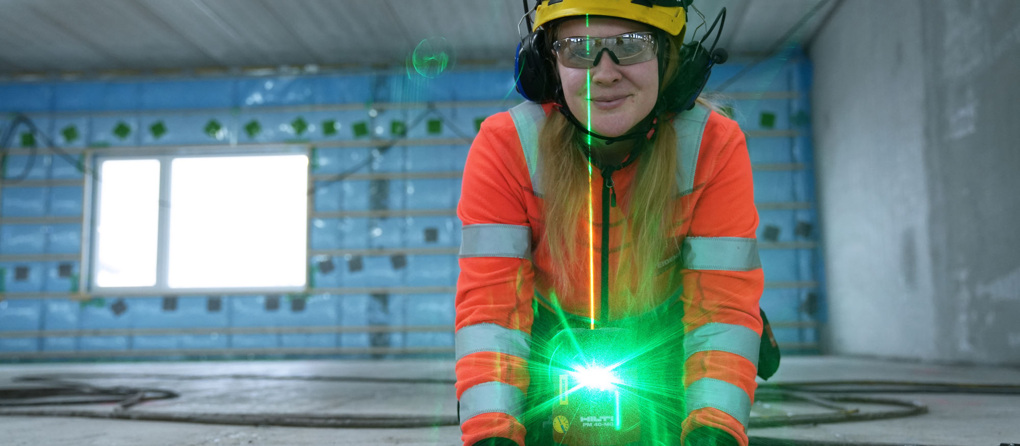 Ung kvinnlig byggarbetare i skyddsutrustning, Stella, står vid grönlysande laserpekare inomhus i ett nybuggnadsprojekt.