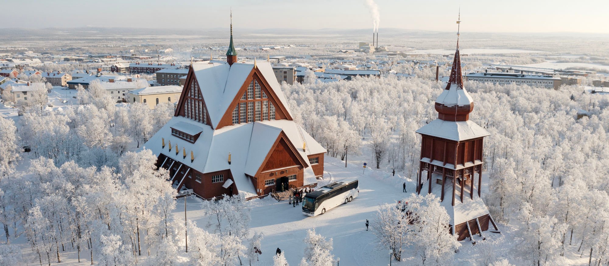 Vinterbild över Kiruna kyrka och klockstapel, två snöklädda röda träbyggnader. 