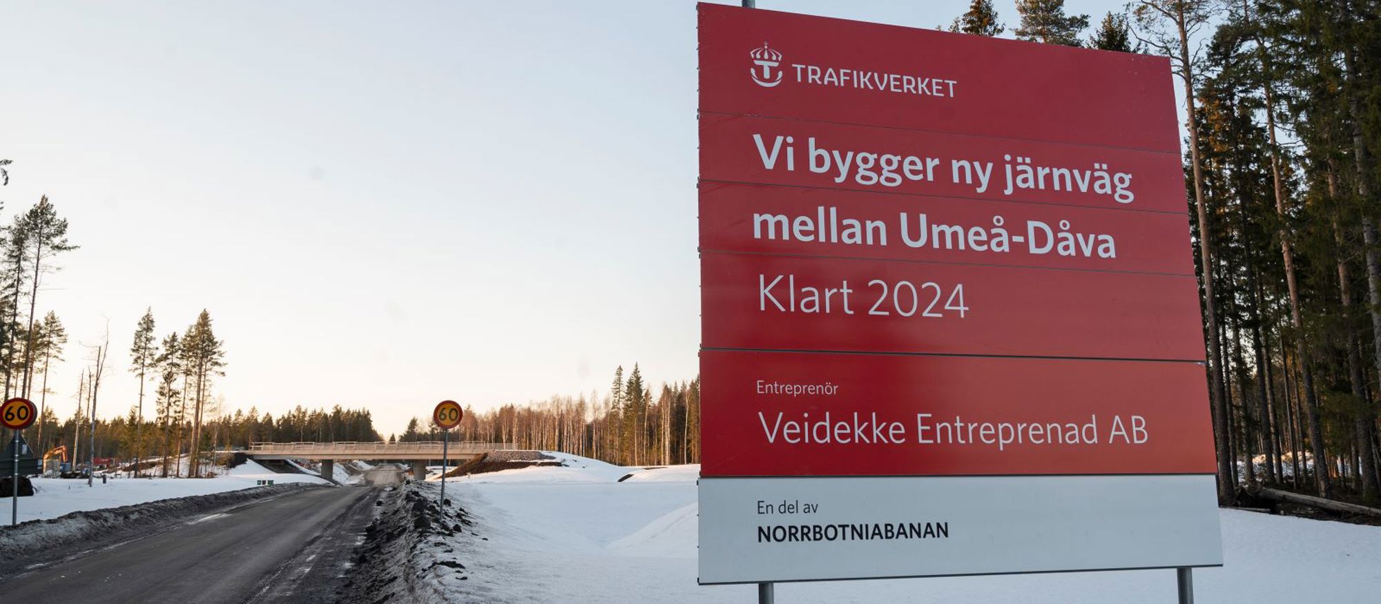 Snöklädd landsväg med viadukt. Förgrunden syns skylt med texten: "Vi bygger ny järnväg mellan Umeå-Dåva. Klart 2024. Trafikverket."