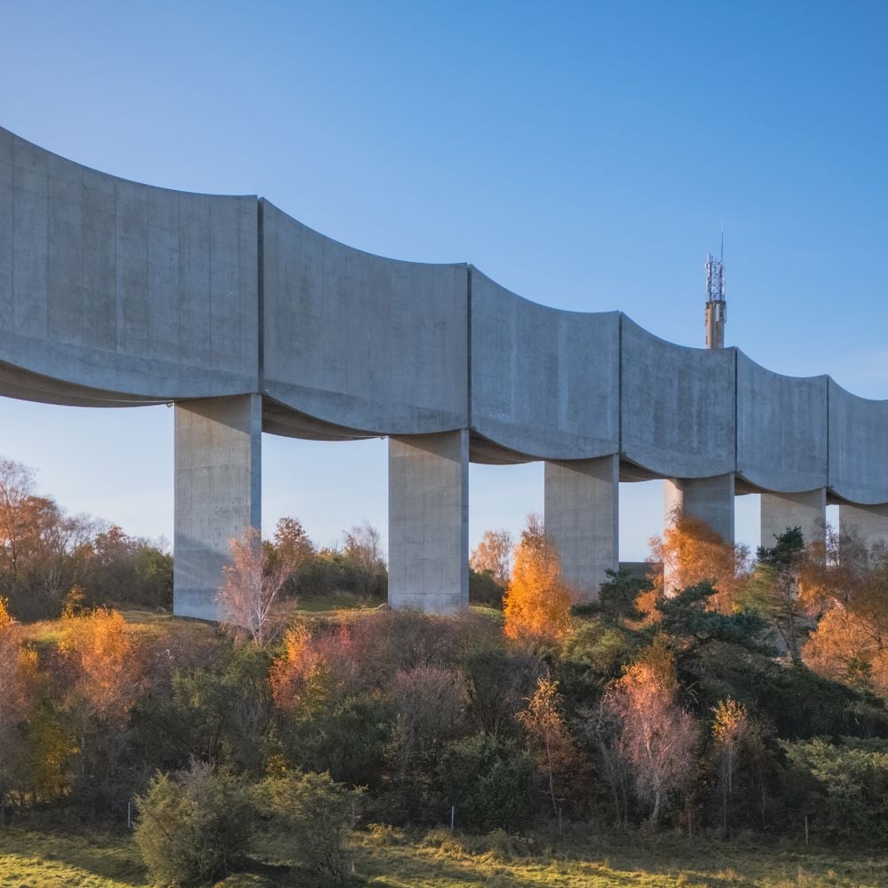 Arkitektbild där Våga vattentorn höjer sig som en våg längs med horisonten. I förgrunden en motorväg framför en bondgård och träd. 