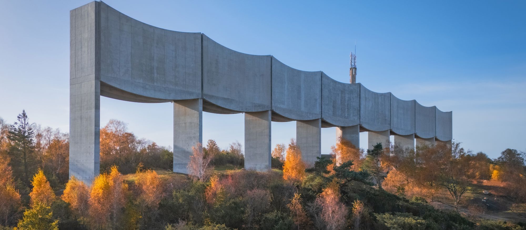 Arkitektbild där Våga vattentorn höjer sig som en våg längs med horisonten. I förgrunden en motorväg framför en bondgård och träd. 