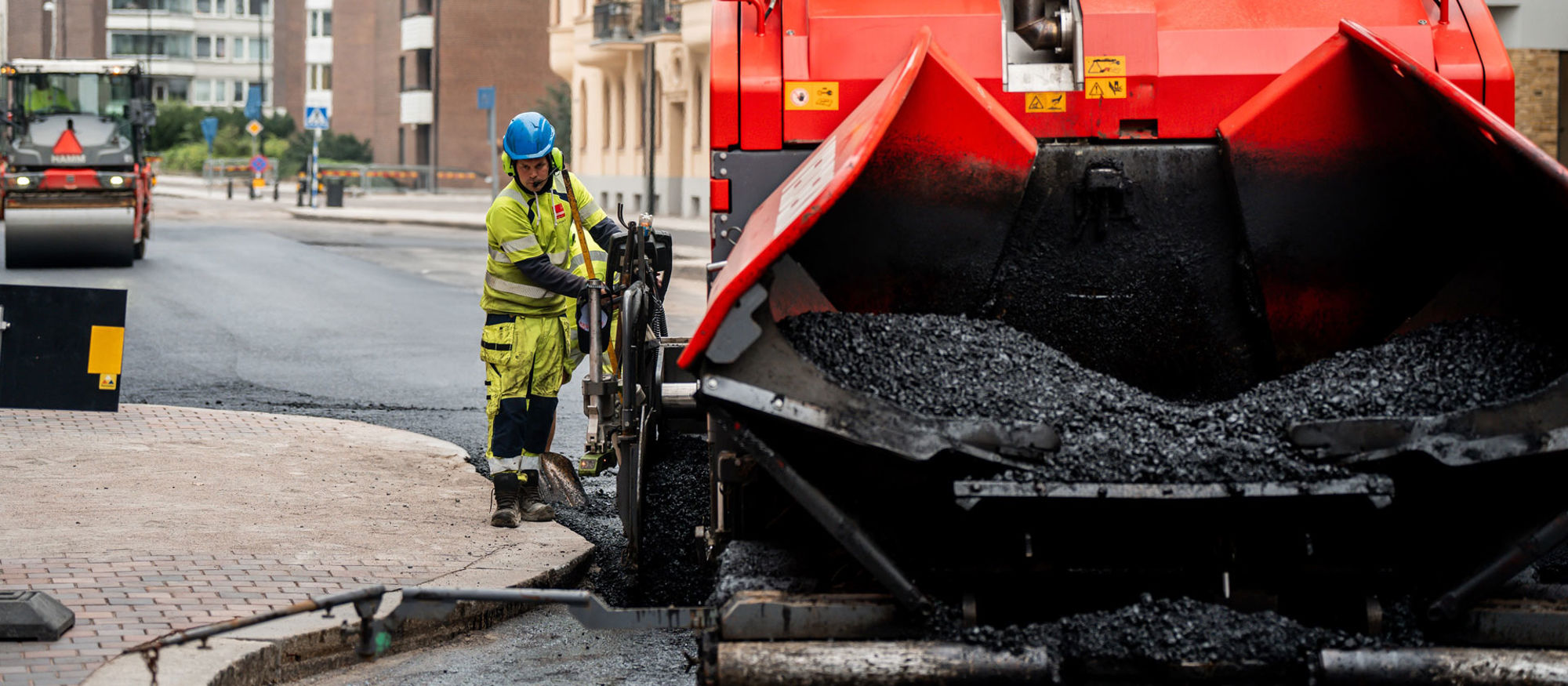 Asfaltsläggning på stadsgata. Skopa med svart asfalt i förgrunden