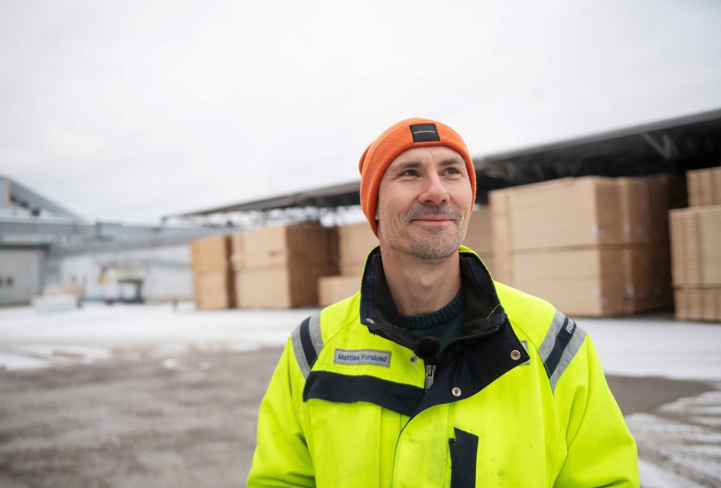 Mattias Forslund. Porträttbild av man med med mössa, klädd i arbetskläder utomhus framför ett virkesupplag. Snö på marken.