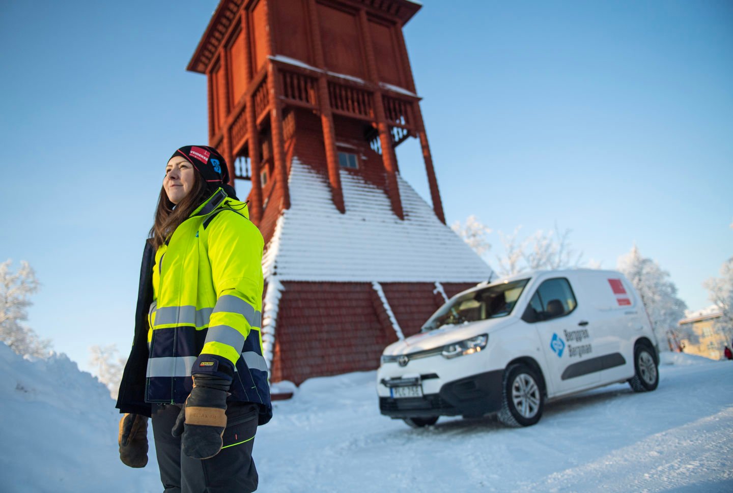 Rött kyrktorn i trä med snö på. Framför står Malin Bergman i varselkläder bredvid en vit skåpbil. 