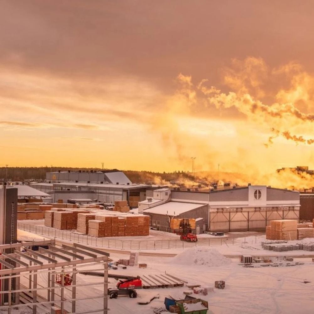 Vintervy över sågverket. Flera industribyygander med rykande skorstenar mot röd-gul himmel. Snö