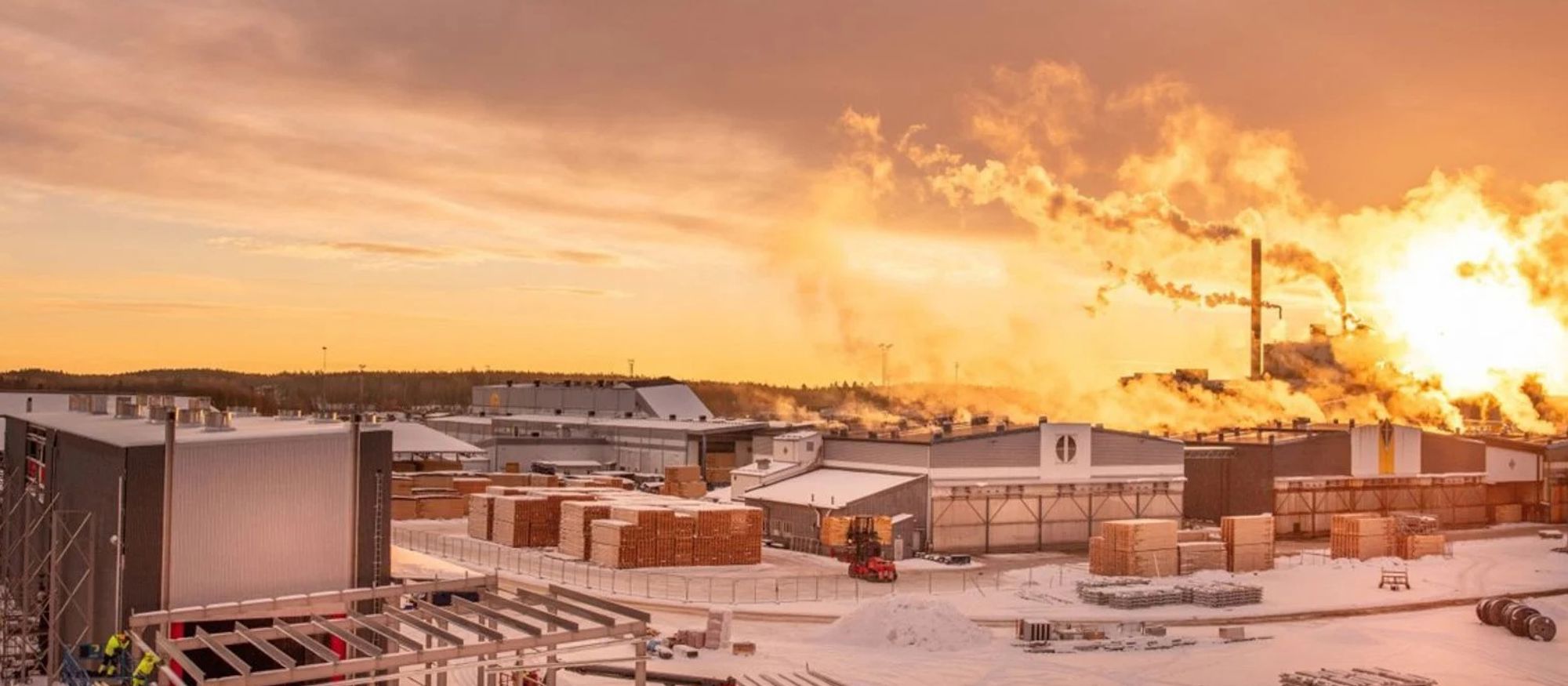 Vintervy över sågverket. Flera industribyygander med rykande skorstenar mot röd-gul himmel. Snö