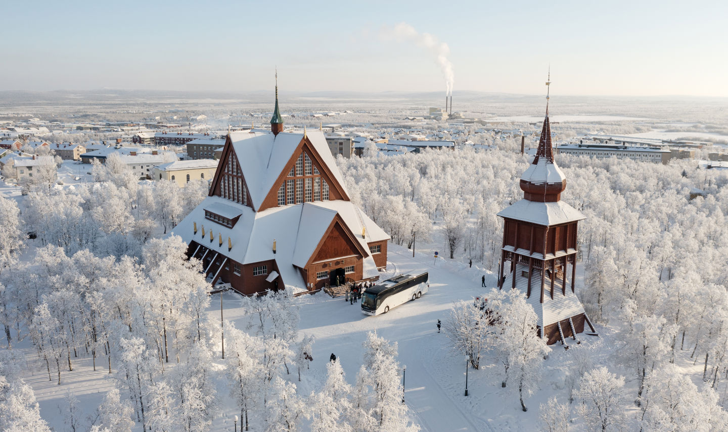 Vinterbild över Kiruna kyrka och klockstapel, två snöklädda röda träbyggnader. 