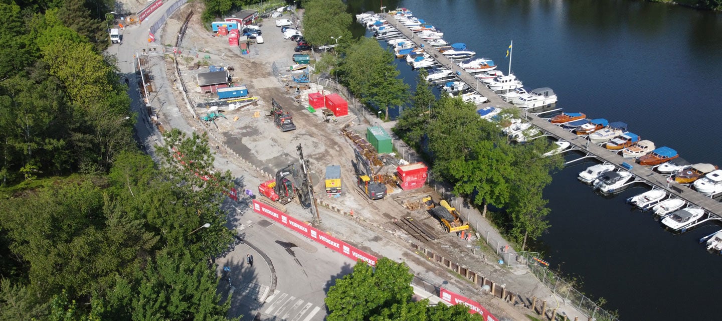 Drönarbild över strandkant med båtar och en byggetablering på sommaren.